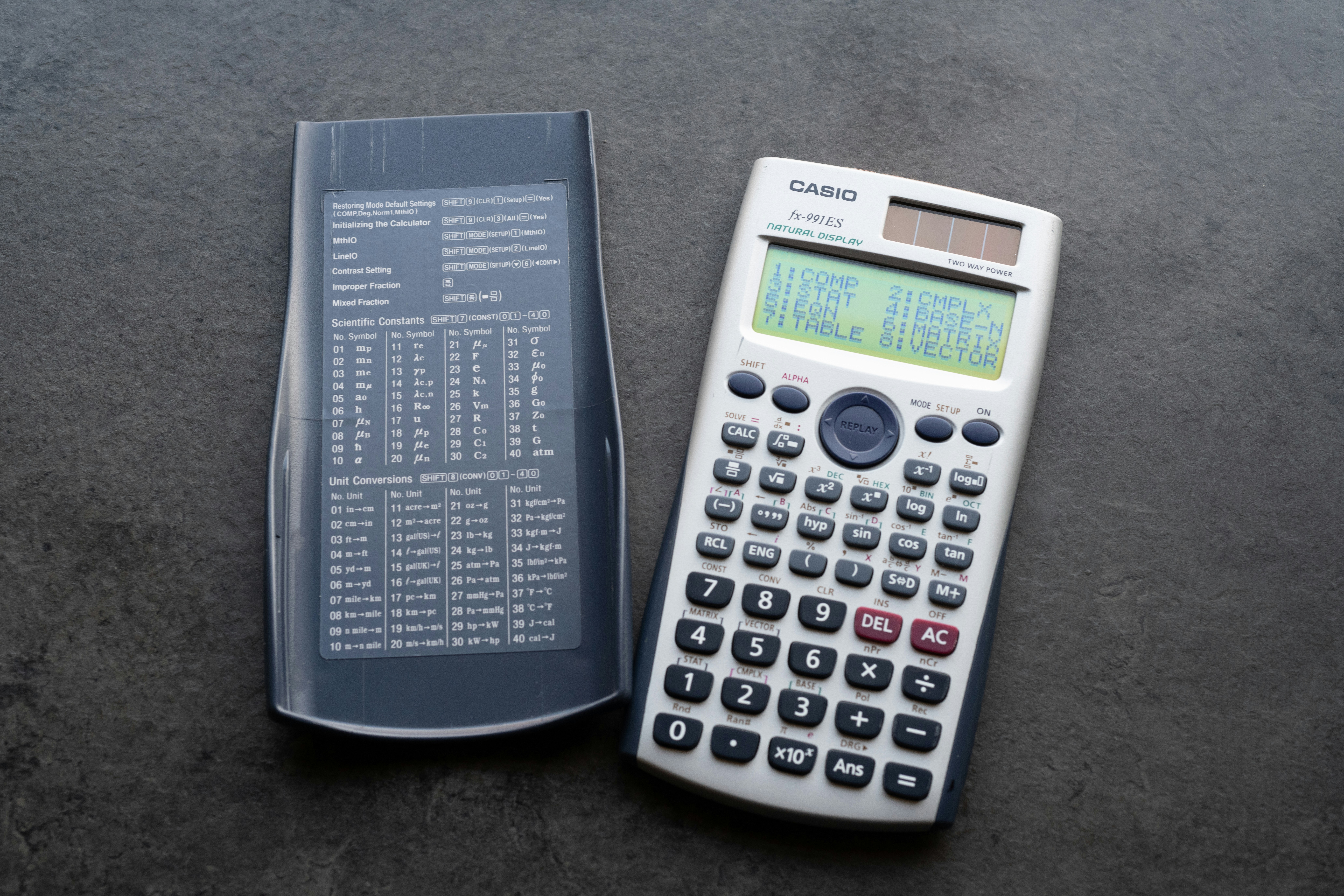 A high-angle, close-up shot features a vintage Casio scientific calculator on a dark gray textured surface. The contrasting colors—off-white and dark gray—create a visually appealing composition. The sharp focus and even lighting highlight the detail of the buttons and displays, offering a nostalgic yet modern aesthetic perfect for tech-related projects.
